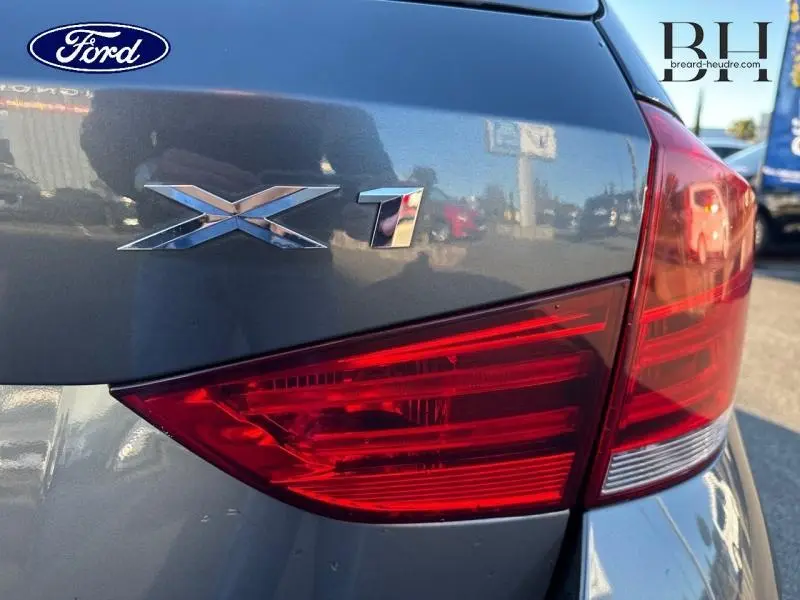 Gros plan sur le feu arrière droit et le logo X1 d’un BMW X1 Mineralgrau, avec reflet de l’environnement urbain.