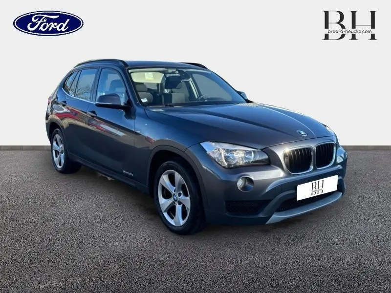BMW X1 sDrive20d gris Mineralgrau vue 3/4 avant droit avec barres de toit noir mat et jantes alu 17 pouces.