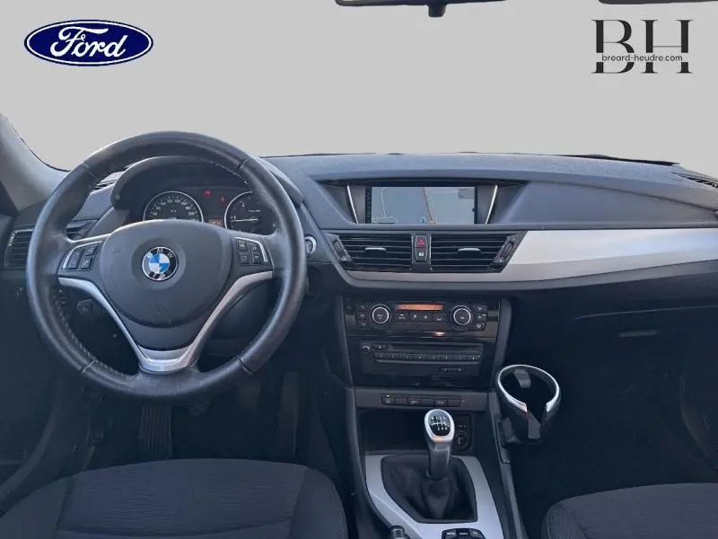 Vue intérieure avant du BMW X1 Mineralgrau 2013, volant cuir avec commandes, écran multifonction et boîte manuelle.