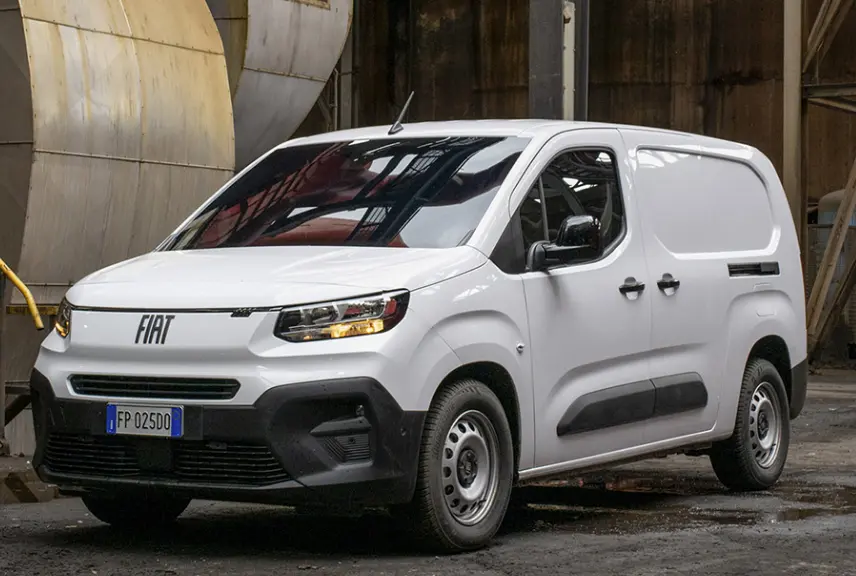 Fourgon Fiat Doblo blanc en 3/4 avant droit, avec calandre noire et feux allumés dans un environnement industriel.