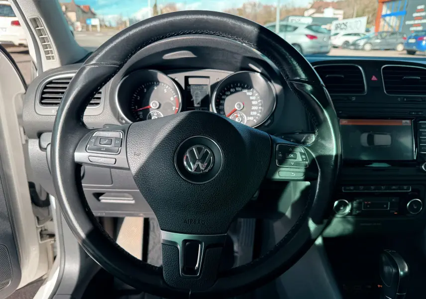 Vue rapprochée du volant en cuir noir d'une Volkswagen Golf blanche de 2012 avec tableau de bord et écran tactile visibles.