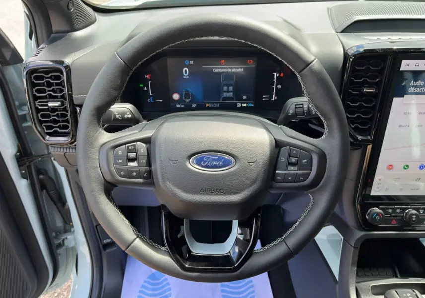 Vue rapprochée du volant cuir multifonctions du Ford Ranger 2.3 PHEV 281 E-AWD avec tableau de bord numérique allumé.