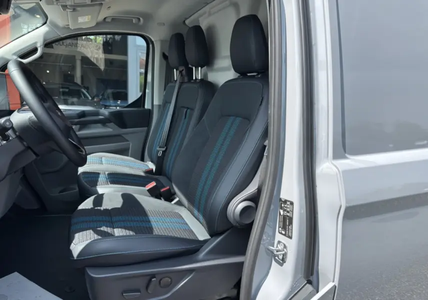 Vue intérieure côté conducteur du Ford Transit Fourgon Custom gris matter, montrant les trois sièges avant en tissu avec détails bleus.