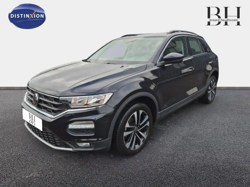 Volkswagen T-Roc gris en 3/4 avant droit, avec jantes alliage et barres de toit visibles.