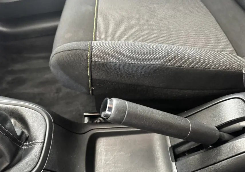 Gros plan sur le frein à main et la console centrale noire, avec siège tissu gris foncé à surpiqûres jaunes visible côté droit.