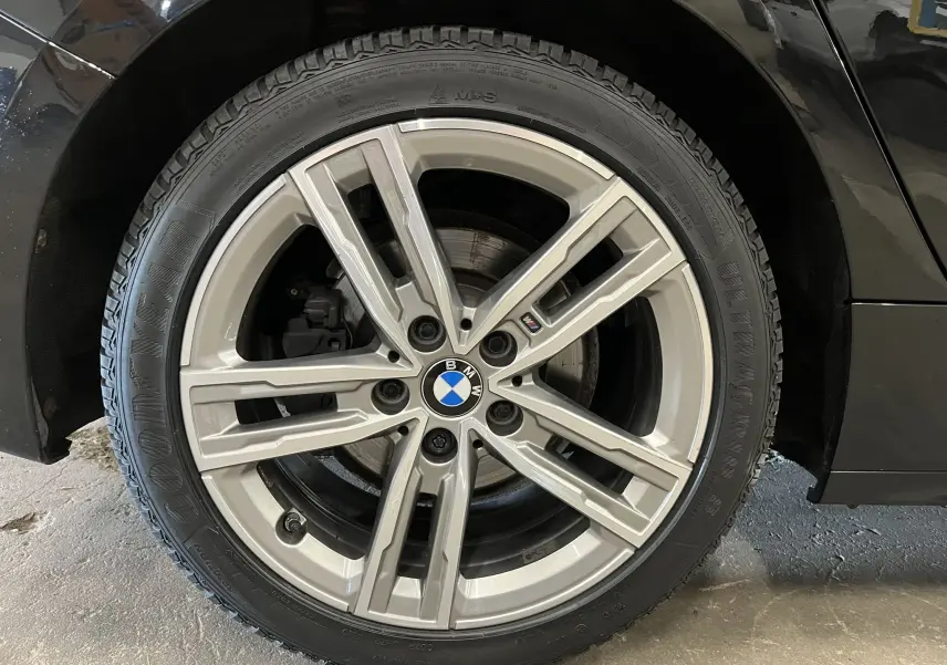 Gros plan sur la roue avant droite d'une BMW Série 1 noire métallisée avec jante alliage M sport et logo BMW central.