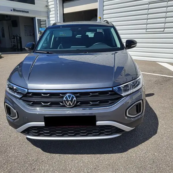 Vue frontale d'un Volkswagen T-Roc gris Indium 2025 garé devant un atelier, avec calandre et phares LED visibles.