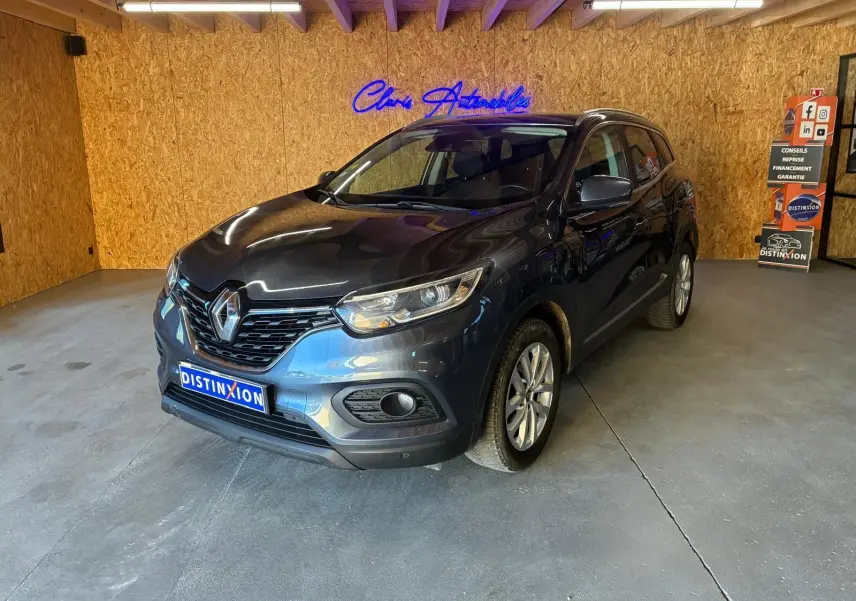 Renault Kadjar gris 2019 en 3/4 avant droit dans un garage avec mur en bois et enseigne lumineuse bleue.