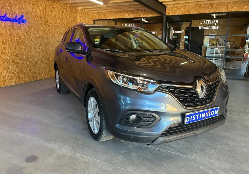 Renault Kadjar gris 3/4 avant droit, SUV compact avec calandre noire et phares LED, stationné en intérieur showroom.