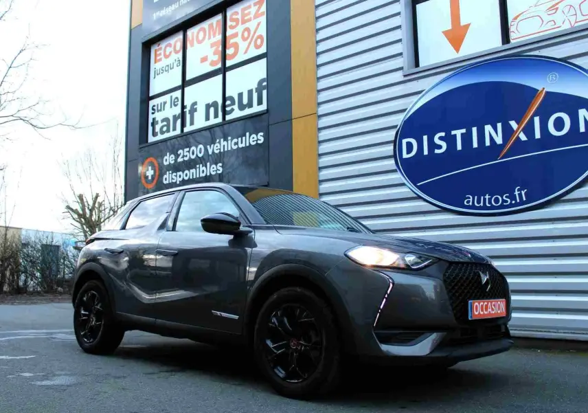 DS3 Crossback gris platinium métal en 3/4 avant droit, avec jantes noires et phares allumés, devant un panneau publicitaire.