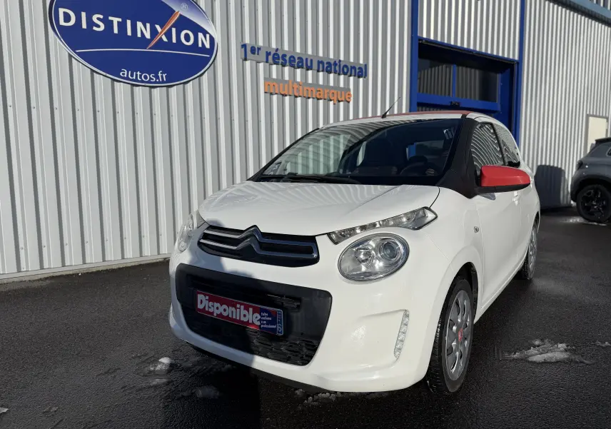 Citroën C1 blanc vue 3/4 avant droit avec rétroviseurs rouges, stationnée devant un bâtiment commercial.