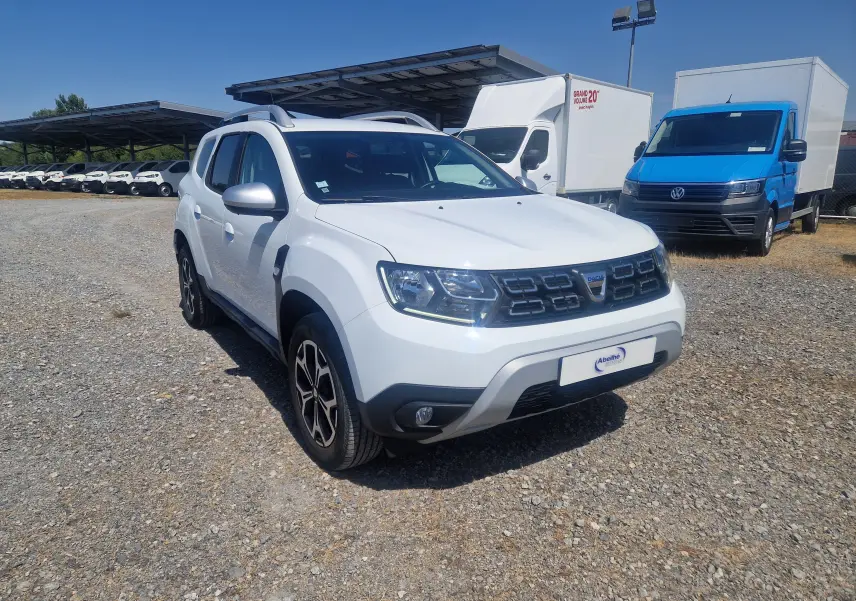 Dacia Duster blanc en 3/4 avant droit, avec calandre noire et jantes alliage, stationné sur un sol gravillonné.