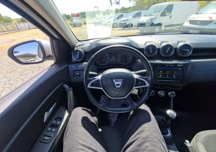 Vue intérieure du poste de conduite du Dacia Duster blanc 2021, avec volant multifonction et levier de vitesse manuel visible.