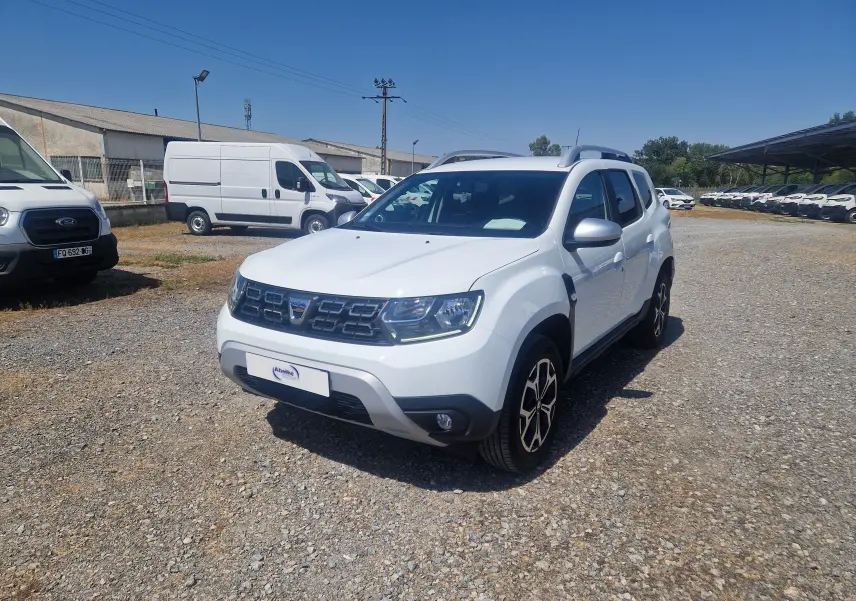 Dacia Duster blanc vue 3/4 avant droit sur parking, avec jantes alliage noires et toit barres de toit grises.