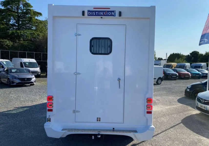 Vue arrière d’un Opel Movano Fourgon blanc avec porte arrière vitrée et feux rouges visibles sur un parking.