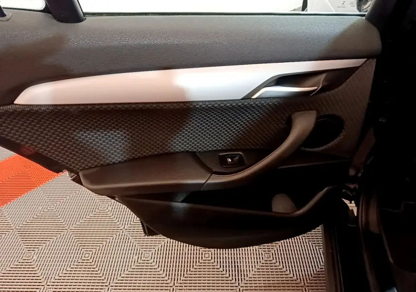 Vue intérieure côté gauche de la porte arrière noire de BMW X2 sDrive18i Lounge avec insert carbone et poignée argentée.