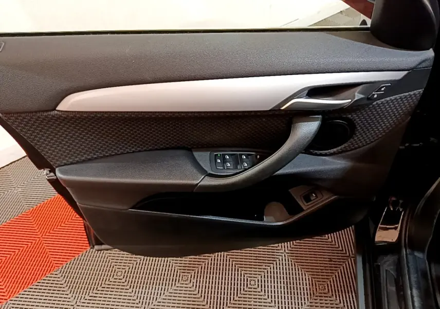 Vue intérieure côté gauche de la porte avant noire de la BMW X2 sDrive18i Lounge avec commandes de vitres et poignée intégrée.