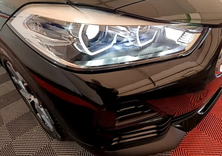 Gros plan sur le phare avant droit allumé d’un BMW X2 sDrive18i Lounge noir, avec détails du pare-chocs et jante visible.