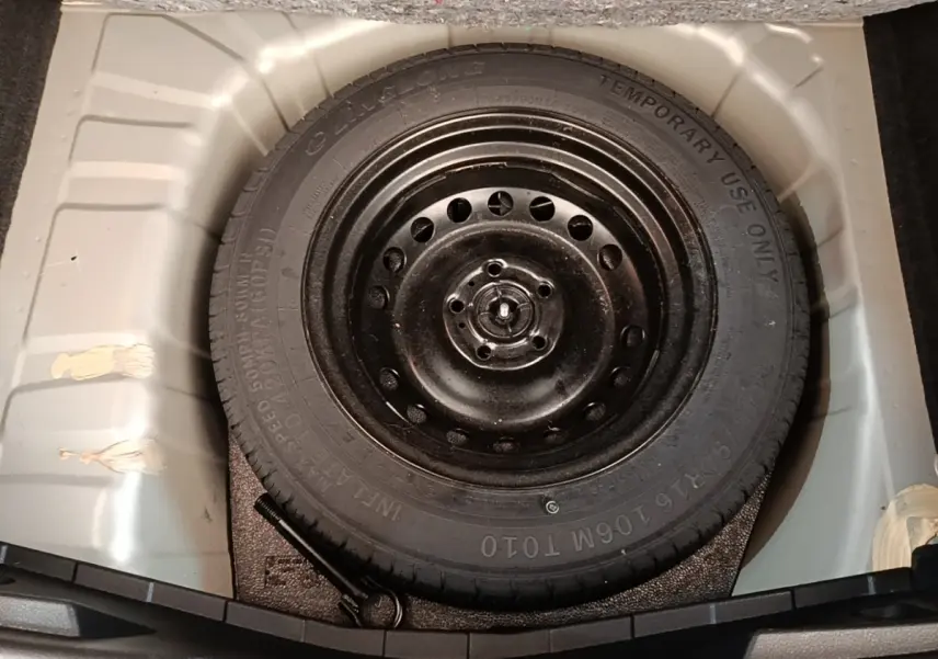 Roue de secours galette noire dans le coffre d’un Nissan Juke gris clair, vue plongeante centrée.