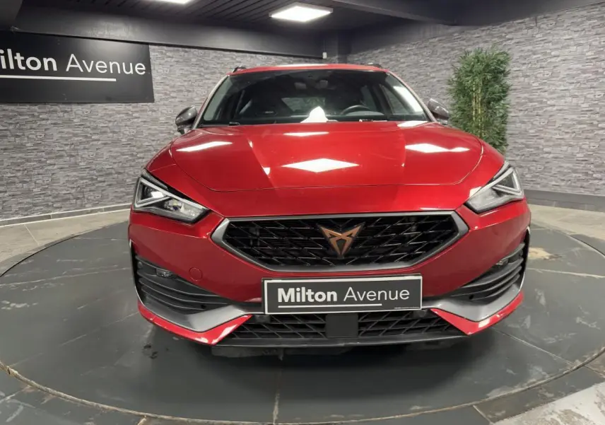 Vue frontale d'une CUPRA Leon Sportstourer rouge Désir avec calandre noire et logo cuivré dans un showroom.