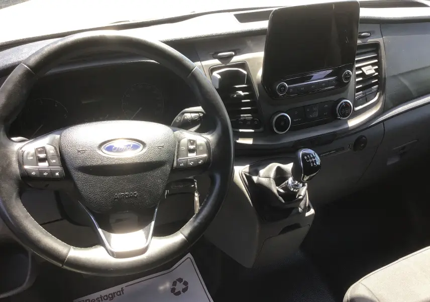 Vue intérieure du poste de conduite du Ford Transit Custom Kombi 2023, volant multifonction et levier de vitesse manuel visibles.