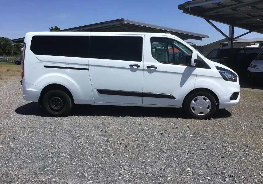 Vue de profil côté gauche d’un Ford Transit Custom Kombi blanc 2023 avec vitres arrière teintées et jantes différentes avant/arrière.