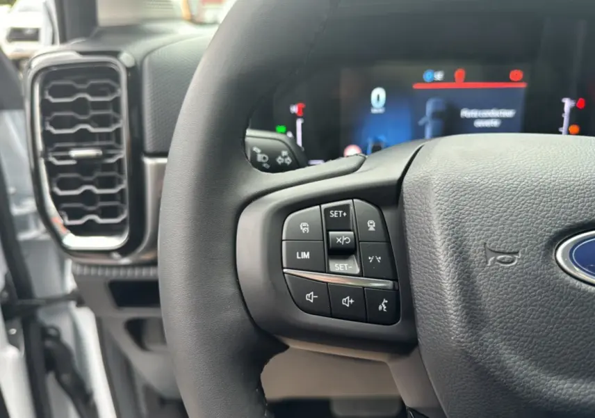 Gros plan sur le volant cuir multifonctions du Ford Ranger blanc glacier, avec commandes intégrées et tableau de bord numérique flou en arrière-plan.