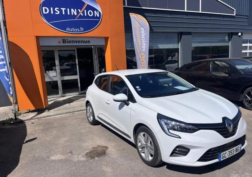 Renault Clio 1.6 E-Tech blanc Quartz vue 3/4 avant droit devant un garage avec enseigne orange.