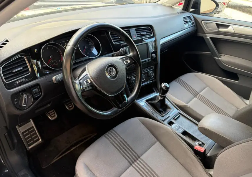 Vue intérieure côté conducteur de la Volkswagen Golf 7 noire, volant cuir multifonctions et levier de vitesse manuel visibles.