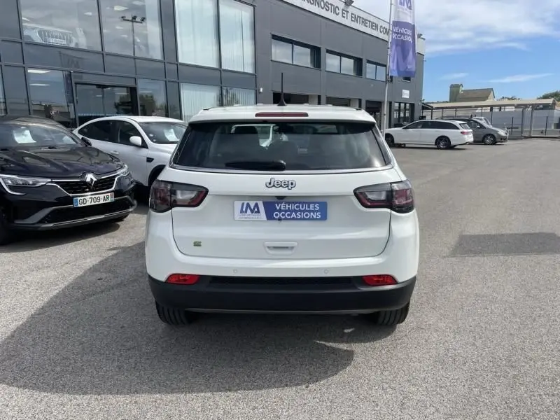 Vue arrière d'un Jeep Compass blanc 2024 hybride essence, avec logo Jeep et plaque "Véhicules Occasions" visible.
