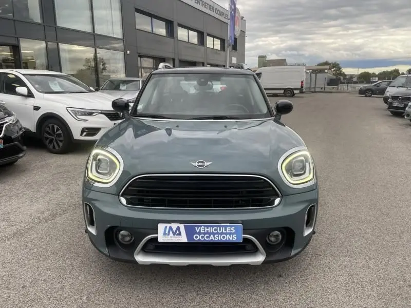 Vue frontale d'une MINI Countryman Cooper 136 gris avec phares allumés sur un parking de véhicules d'occasion.
