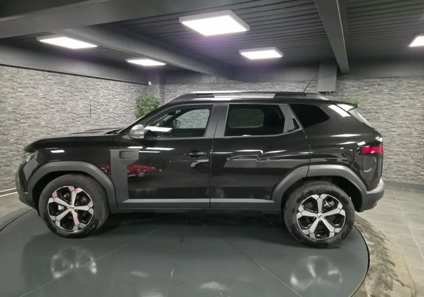 Profil droit du Dacia Duster 1.6 Hybrid 2025 noir nacré avec jantes alliage 18 pouces et barres de toit noires.