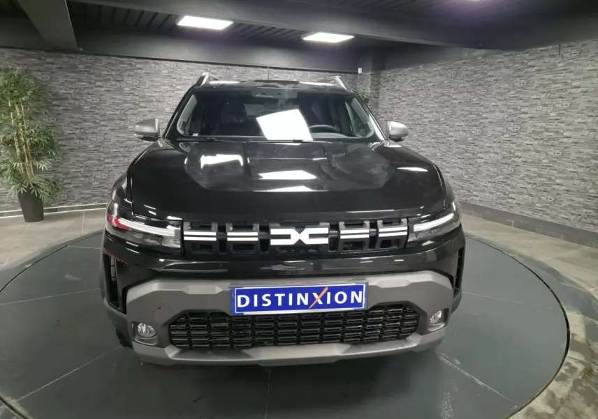 Vue avant d'un Dacia Duster 1.6 Hybrid noir nacré avec calandre noire et bouclier gris, en showroom intérieur.