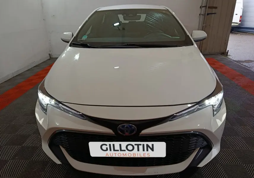 Vue avant d'une Toyota Corolla hybride blanche 2022 avec calandre noire et phares LED allumés dans un garage.