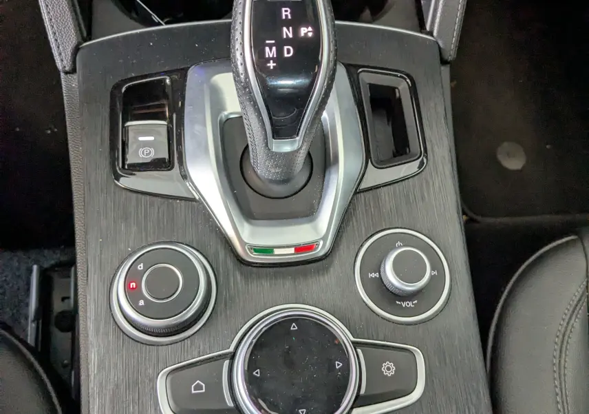 Vue plongeante sur la console centrale noire de l'Alfa Romeo Stelvio 2023, mettant en valeur le levier de vitesses automatique et les commandes multimédia.