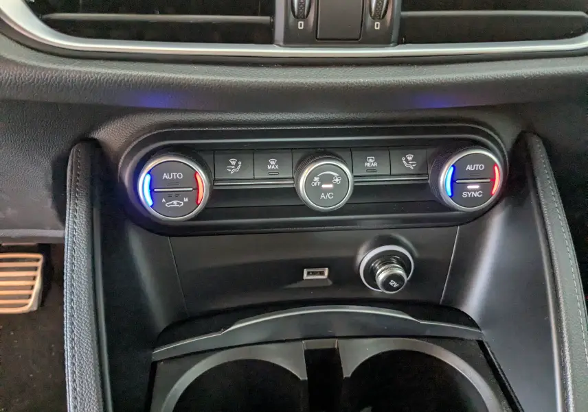 Vue rapprochée de la console centrale de l'Alfa Romeo Stelvio noir, montrant les commandes de climatisation automatique avec éclairage bleu et rouge.