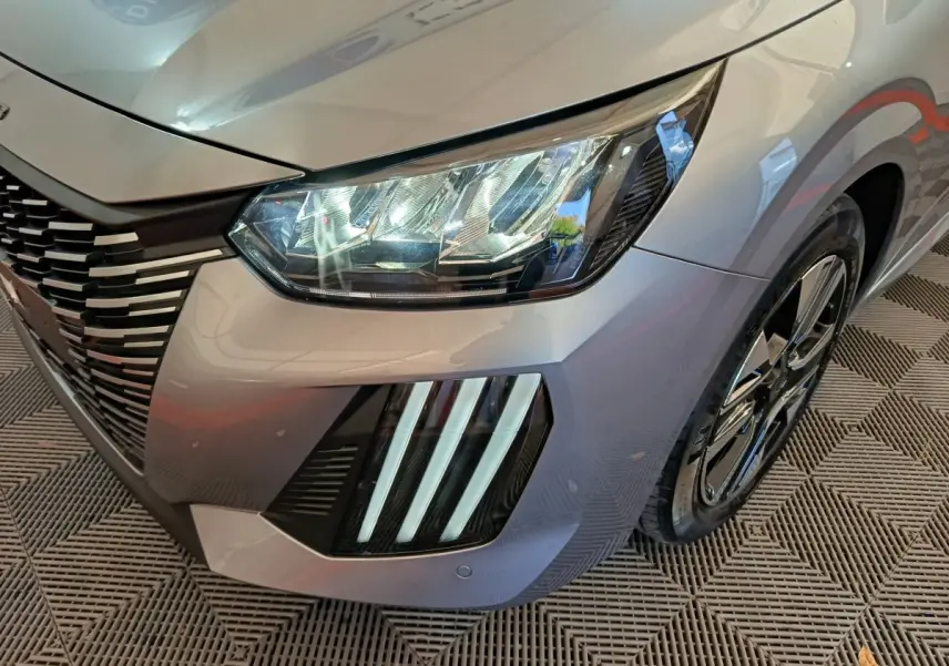 Gros plan sur l'avant droit gris clair du Peugeot 208 2025, mettant en valeur les phares LED et la calandre distinctive.
