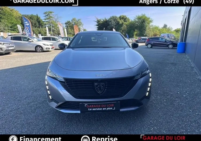 Vue avant frontale d'une Peugeot 308 gris clair 2023 avec feux LED verticaux allumés sur un parking extérieur.