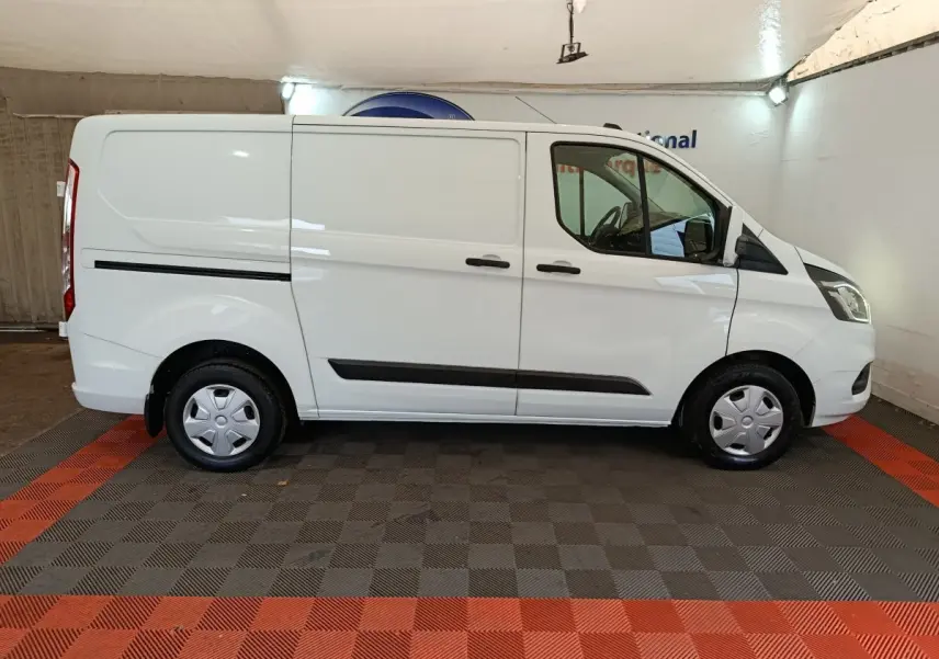 Vue de profil côté droit d’un Ford Transit Custom blanc, fourgon utilitaire avec jantes acier et protections noires sur les portières.