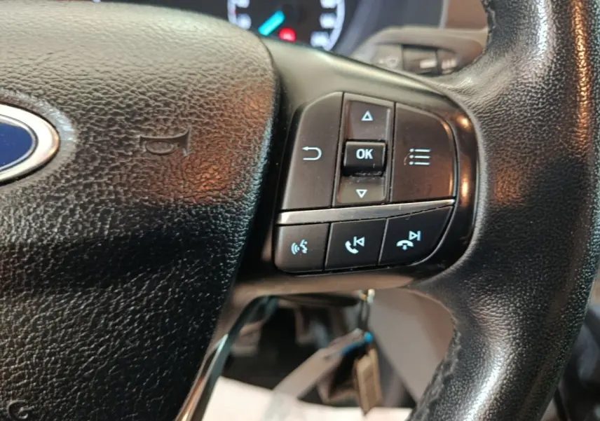 Gros plan sur le volant noir du Ford Transit Custom avec commandes multifonctions et logo Ford visible.