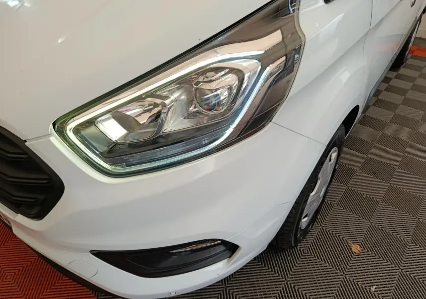 Gros plan sur le phare avant droit du Ford Transit Custom blanc, avec partie de la roue et pare-chocs visibles.