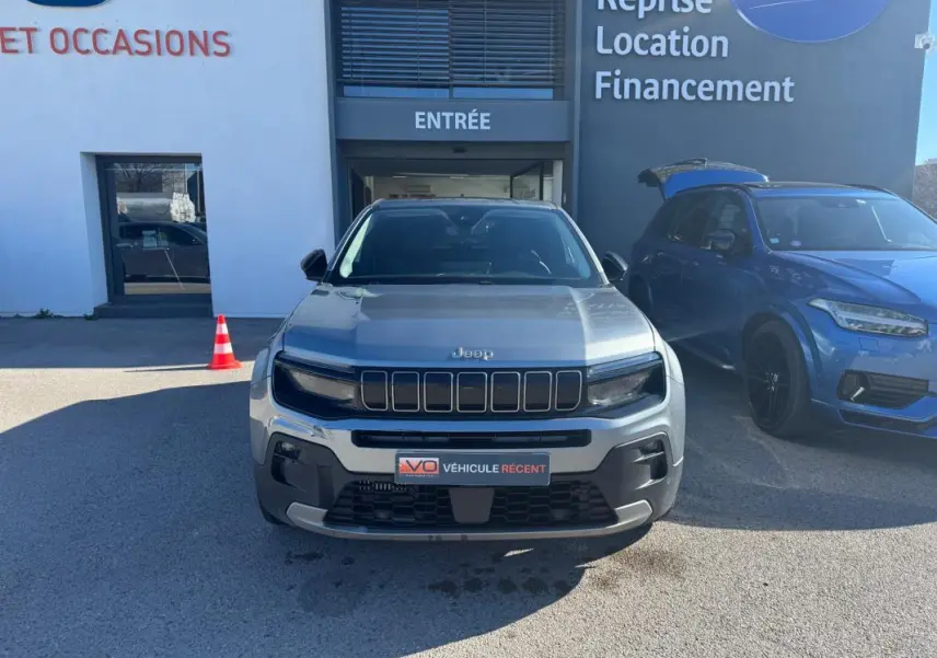 Vue de face d'un Jeep Avenger gris 2025 garé devant un bâtiment avec calandre noire distinctive et phares allumés.
