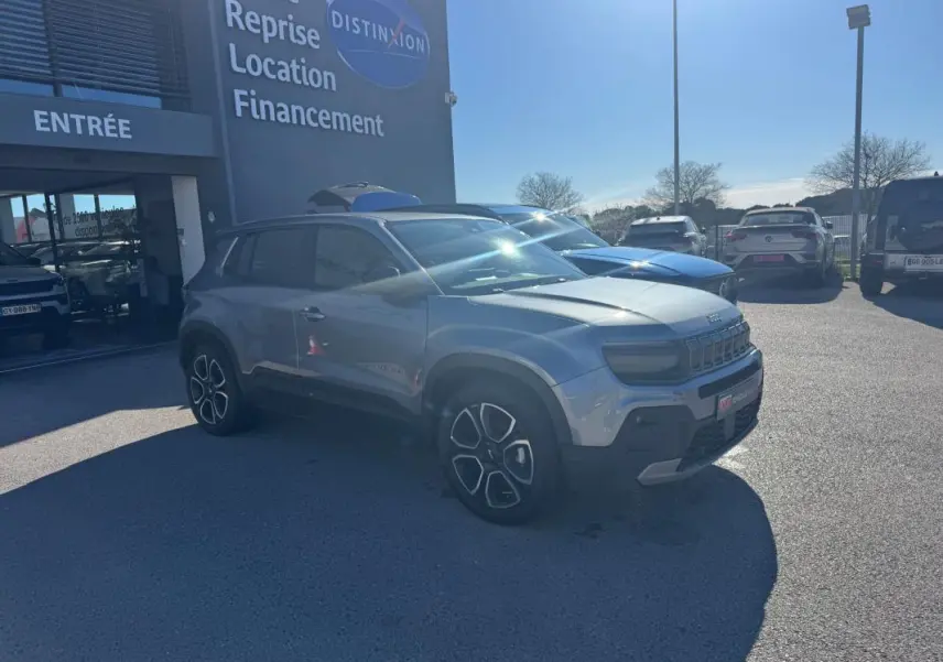 Jeep Avenger gris en 3/4 avant droit, avec jantes alliage 18 pouces, stationné devant un bâtiment commercial.