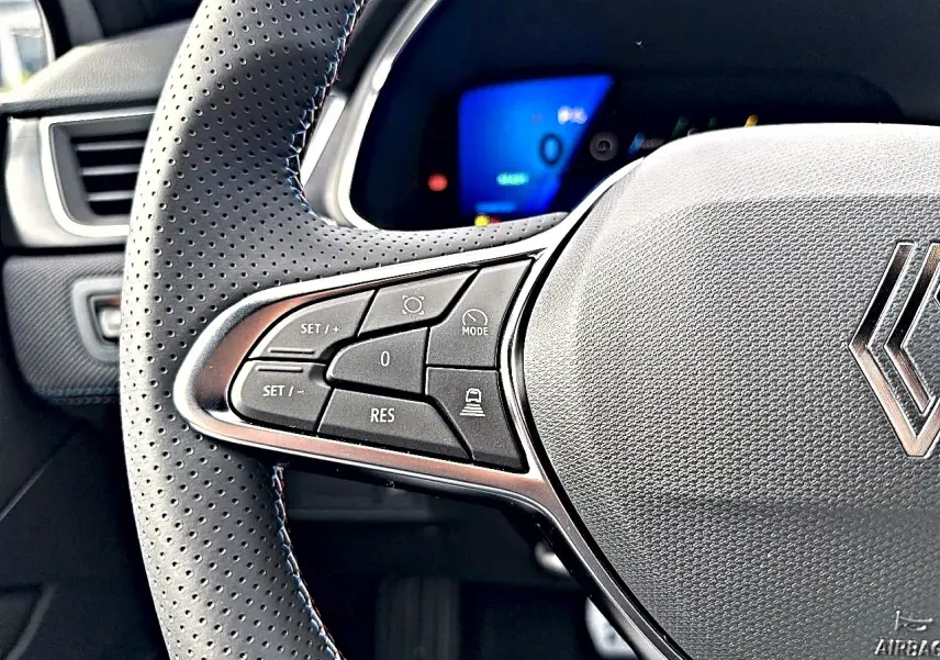 Gros plan sur le côté gauche du volant perforé gris avec commandes multifonctions et logo Renault central.