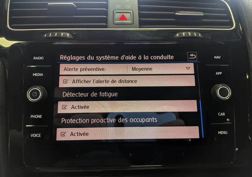 Vue rapprochée de l'écran tactile central noir du Volkswagen Golf VII blanc, affichant les réglages d'aide à la conduite.