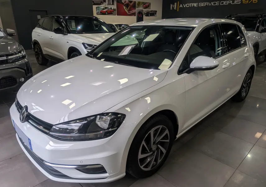 Vue 3/4 avant droite d'une Volkswagen Golf VII TSI 125 CH Sound blanche Pure en showroom avec jantes alliage et vitres teintées.