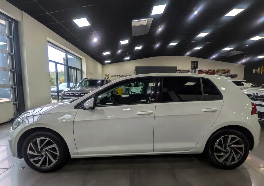 Profil côté gauche d'une Volkswagen Golf VII TSI 125 CH Sound blanche, avec jantes alliage et vitres teintées en showroom.