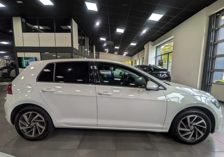 Profil côté gauche d'une Volkswagen Golf VII TSI blanche, avec jantes alliage et vitres teintées en showroom.
