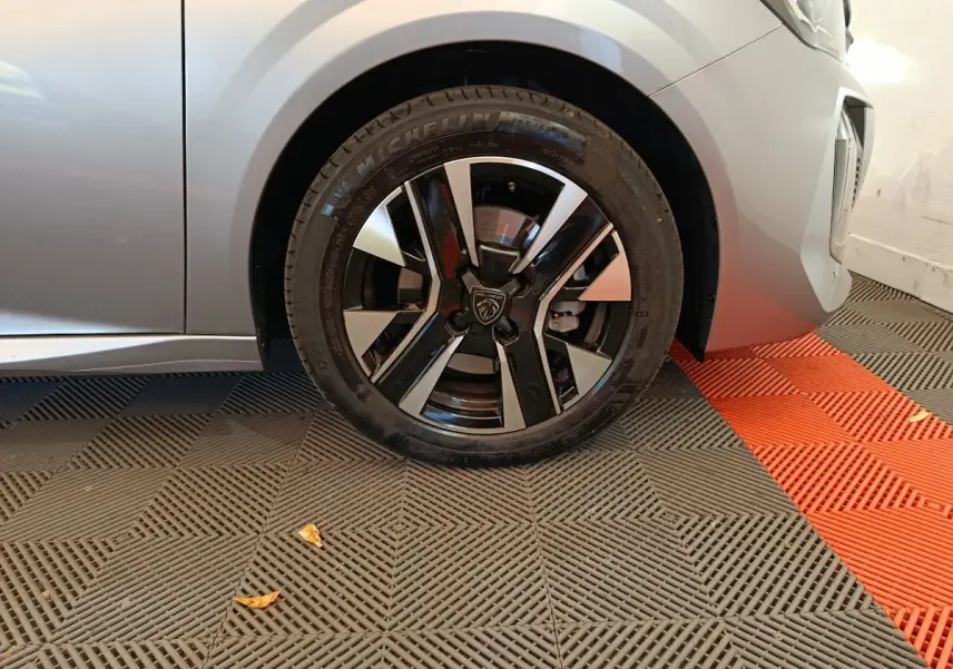 Gros plan sur la roue avant gauche d'une Peugeot 208 gris clair, avec jante noire et argentée au design moderne.
