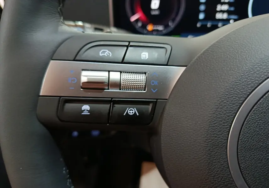 Gros plan sur les commandes multifonctions à gauche du volant du Hyundai Tucson 1.6 Hybrid 239 gris foncé, tableau de bord numérique en arrière-plan.